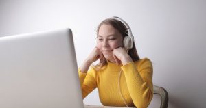 Woman in a yellow sweater wearing headphones, resting her chin in her hands and smiling at her laptop