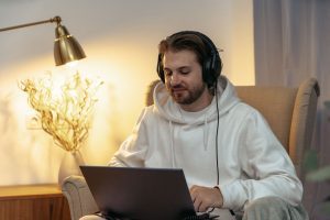 Man wearing headphones using a laptop in an armchair at home, with a lamp on beside him
