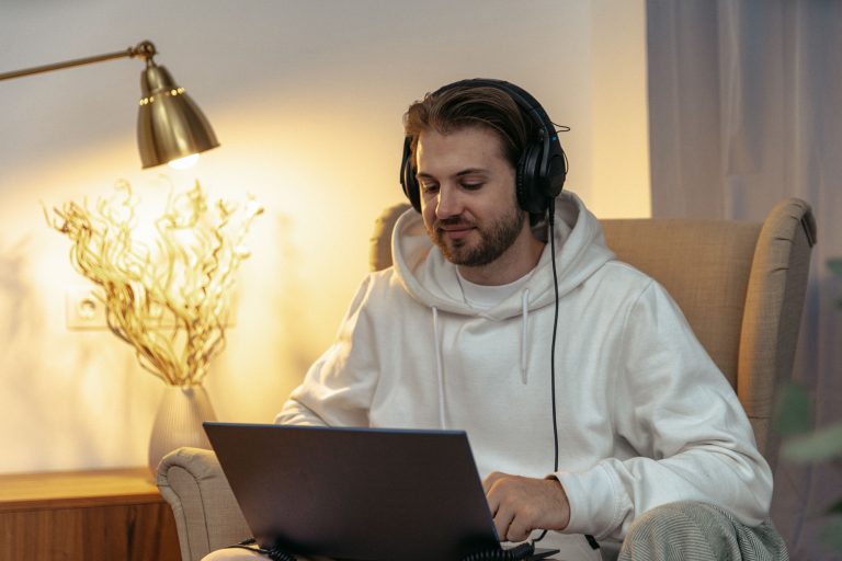 Man wearing headphones using a laptop in an armchair at home, with a lamp on beside him